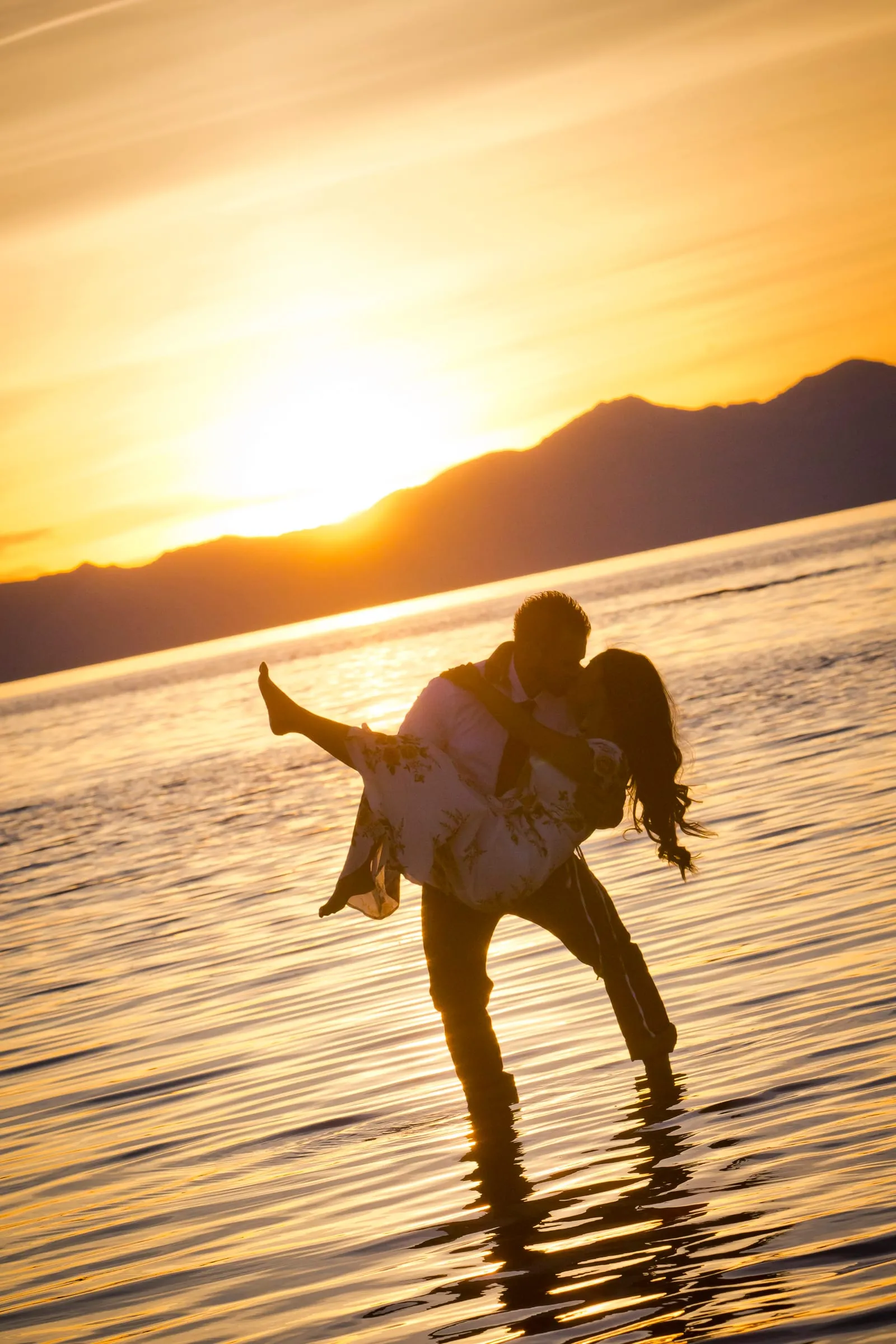Man dipping his fiancée in shallow water as the sun sets behind the mountains