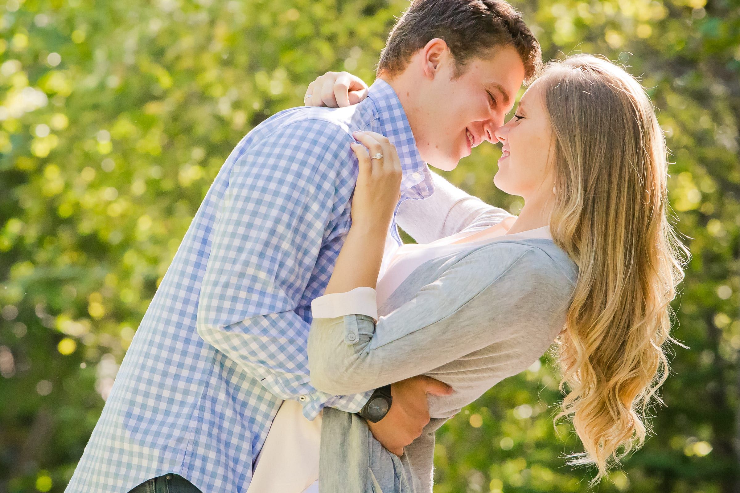 Engaged couple leaning in nose to nose in a sunny, leafy park